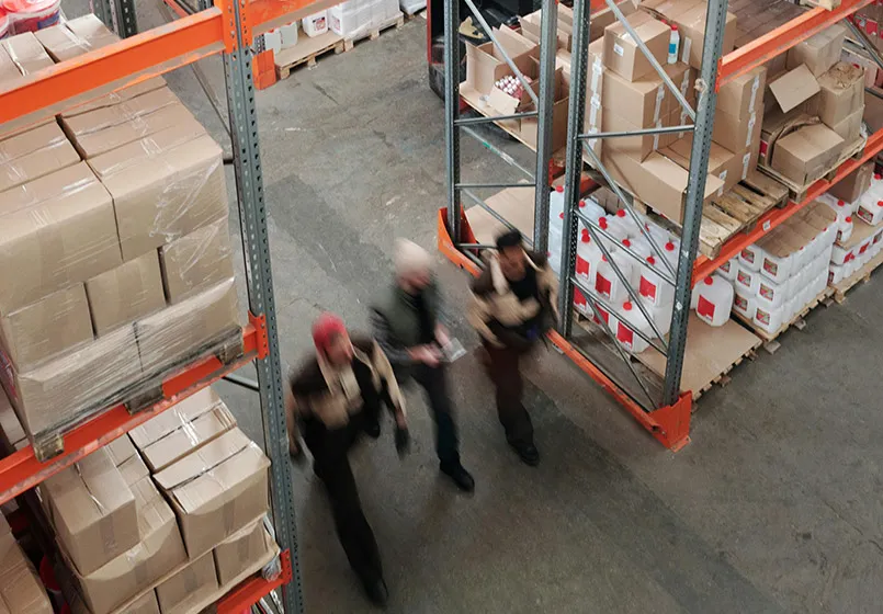 Drie werknemers lopen door een magazijn met stellingen vol dozen en jerrycans.